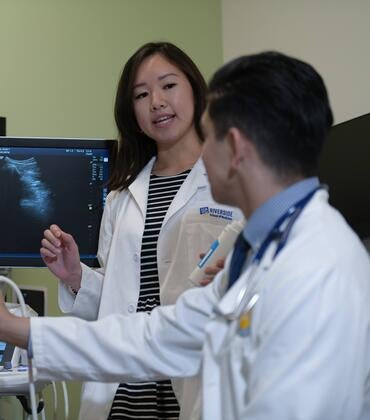 A medical student teaches another student how to use an ultrasound device while a standardized patient looks on.