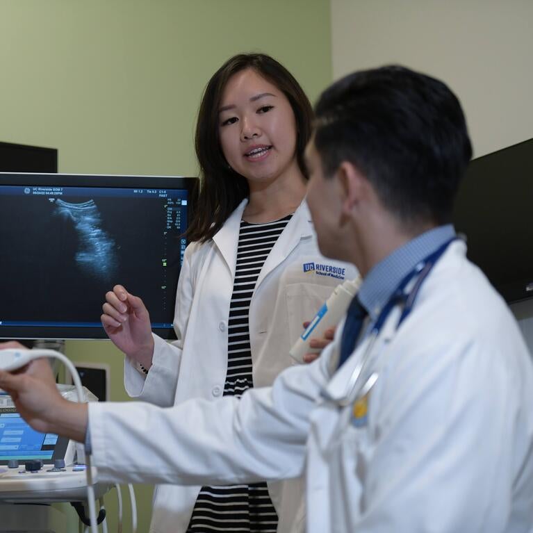 A medical student teaches another student how to use an ultrasound device while a standardized patient looks on.